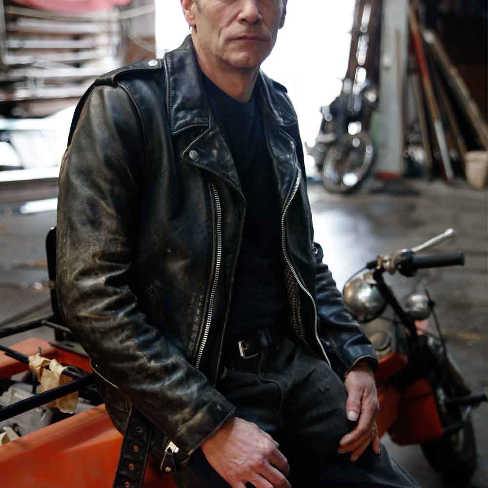 A man wearing a distressed black leather motorcycle jacket sits on an orange vehicle in a workshop setting with a motorcycle visible behind him.