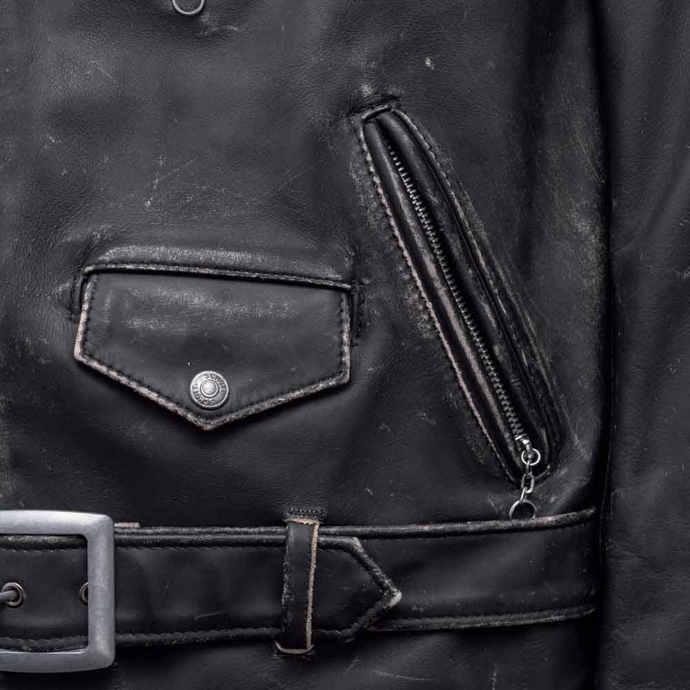 Close-up of a black leather jacket showing a flap pocket with a silver snap button, a zippered pocket, and part of a matching leather belt with a silver buckle.