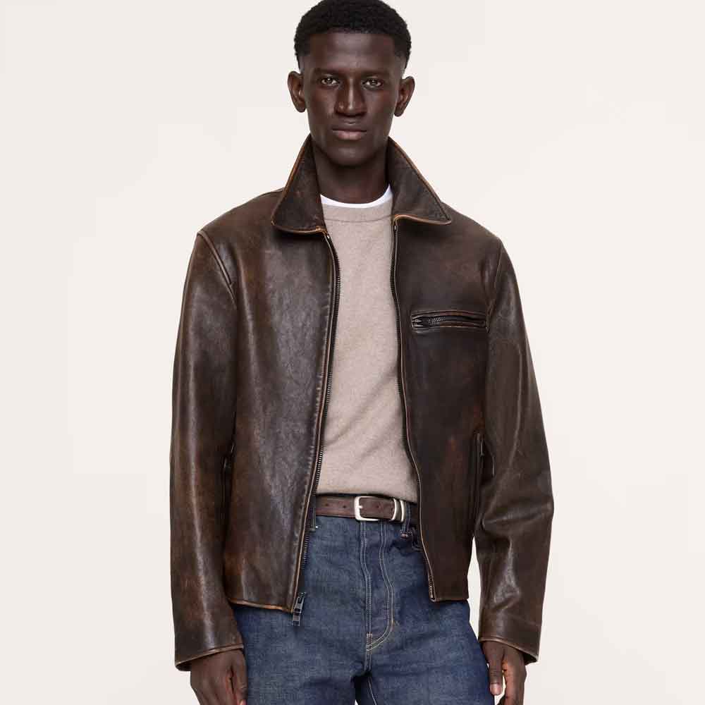 A male model wearing a dark brown distressed leather aviator jacket over a beige crewneck sweater, blue jeans, and a brown leather belt, photographed against a solid off-white studio background.
