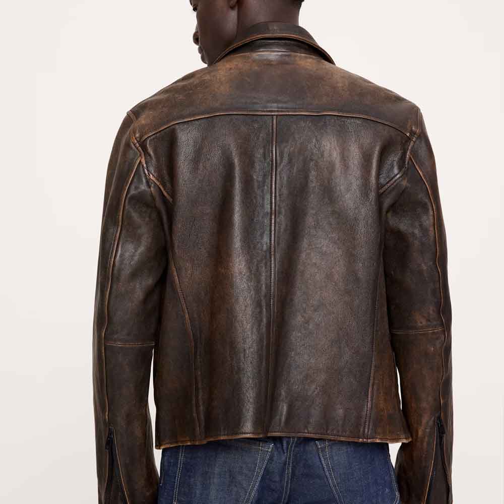 Rear view of a model wearing a dark brown distressed leather jacket with contrast stitching, paired with blue jeans, against a plain off-white background.