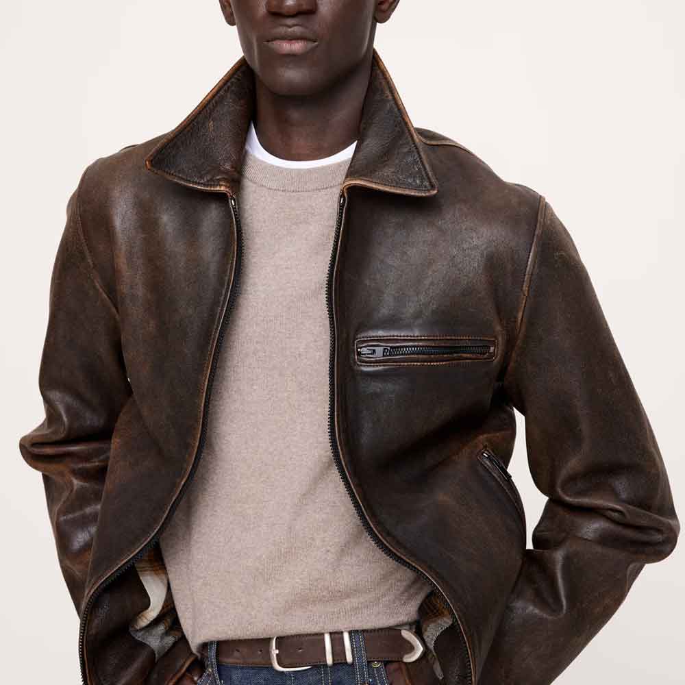 A man wearing a dark brown distressed leather aviator-style jacket over a beige crewneck sweater and blue jeans, photographed against a clean off-white studio background.