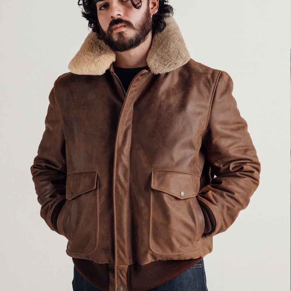A man wearing a brown leather aviator-style jacket with a thick beige shearling collar, standing against a plain light background.