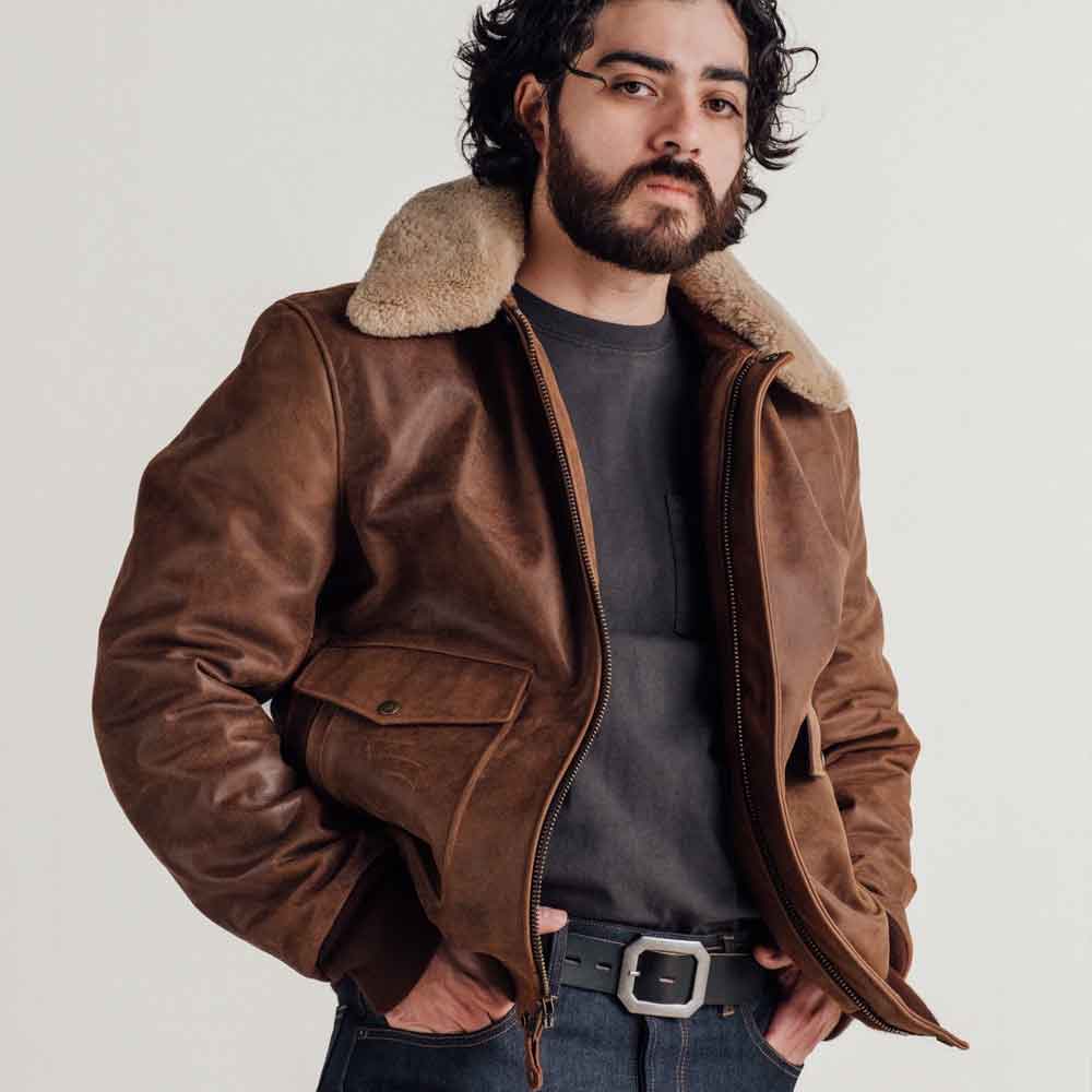 A man wearing a brown leather aviator-style jacket with a thick beige shearling-style collar, layered over a dark gray t-shirt and blue jeans.
