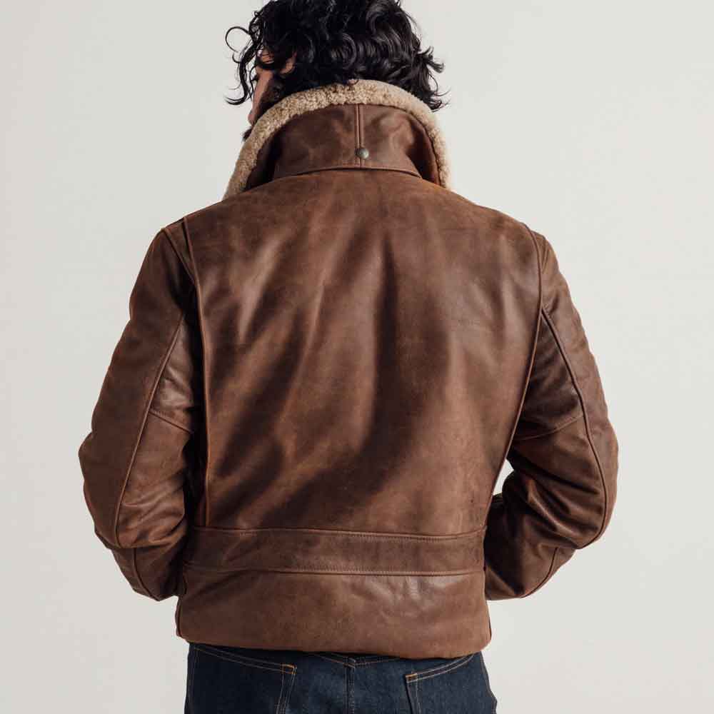 A man viewed from behind wearing a brown leather aviator-style jacket with a beige shearling lining on the collar, paired with dark blue jeans against a plain light gray background.