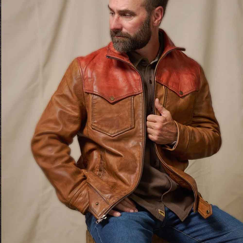 A man wearing a brown leather jacket with reddish-orange shoulder panels, blue jeans, and a dark shirt, posed against a neutral beige backdrop.
