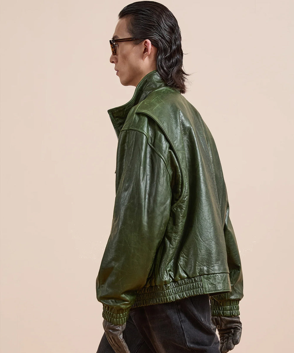 A man in profile wearing a glossy olive green leather jacket, black pants, and sunglasses against a solid beige background.