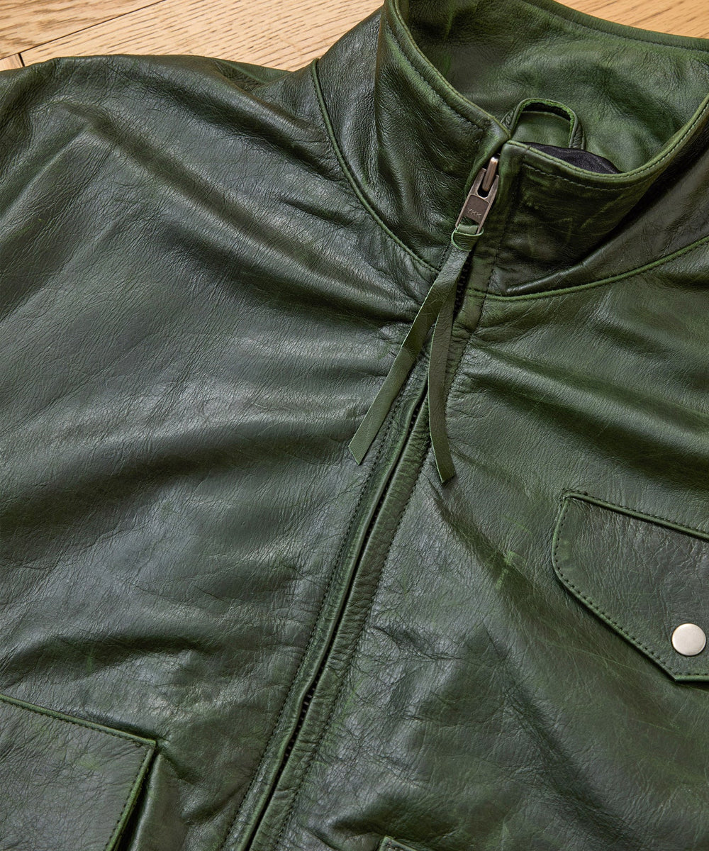 Close-up of an olive green leather jacket with a high collar, front zipper, and a flap pocket with a silver snap button, laid flat on a light wood surface.