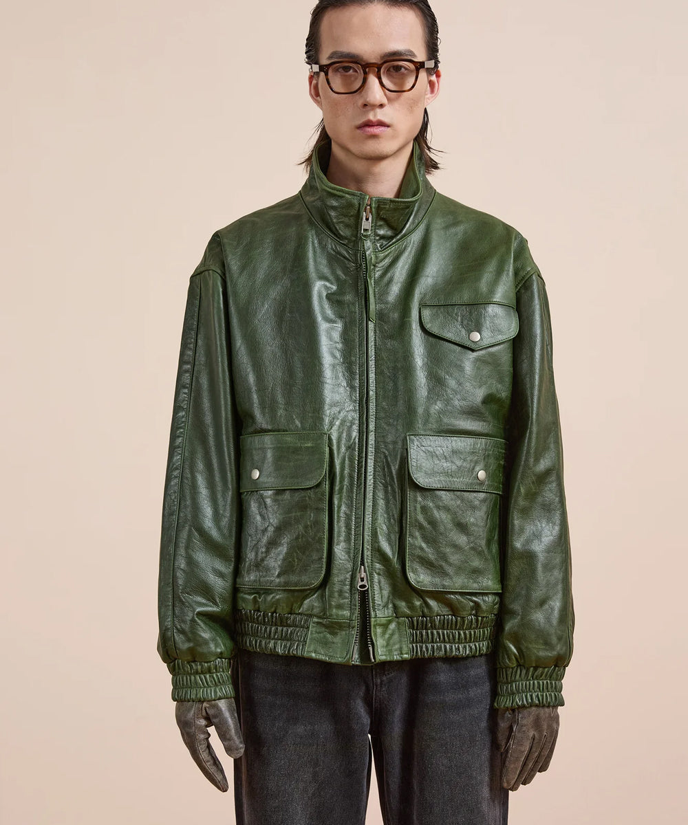 A model wearing a dark green leather utility jacket with a high collar, front zipper, and four front flap pockets, styled with dark pants and gloves against a solid beige background.